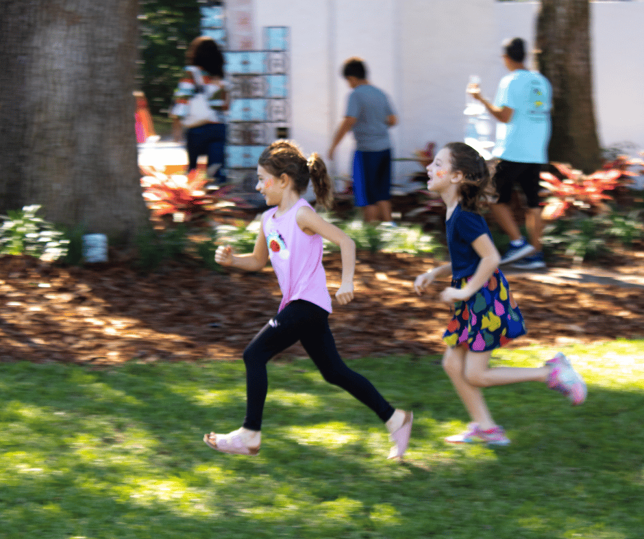 Two young white children run, laughing, through the A&H garden, other families a blur in the background