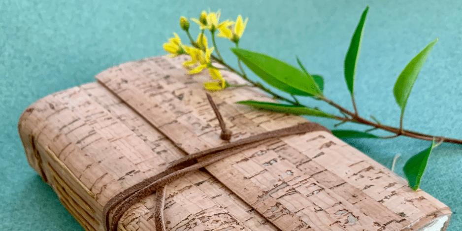 Hand bound book made with cork materials placed on a teal background and accompanied by flowers.
