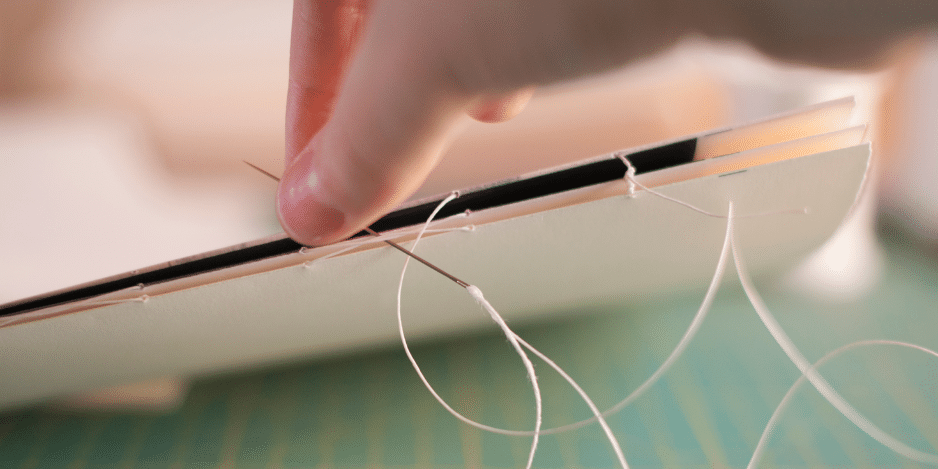 Hand holding a thread and needle, binding paper together for a book.