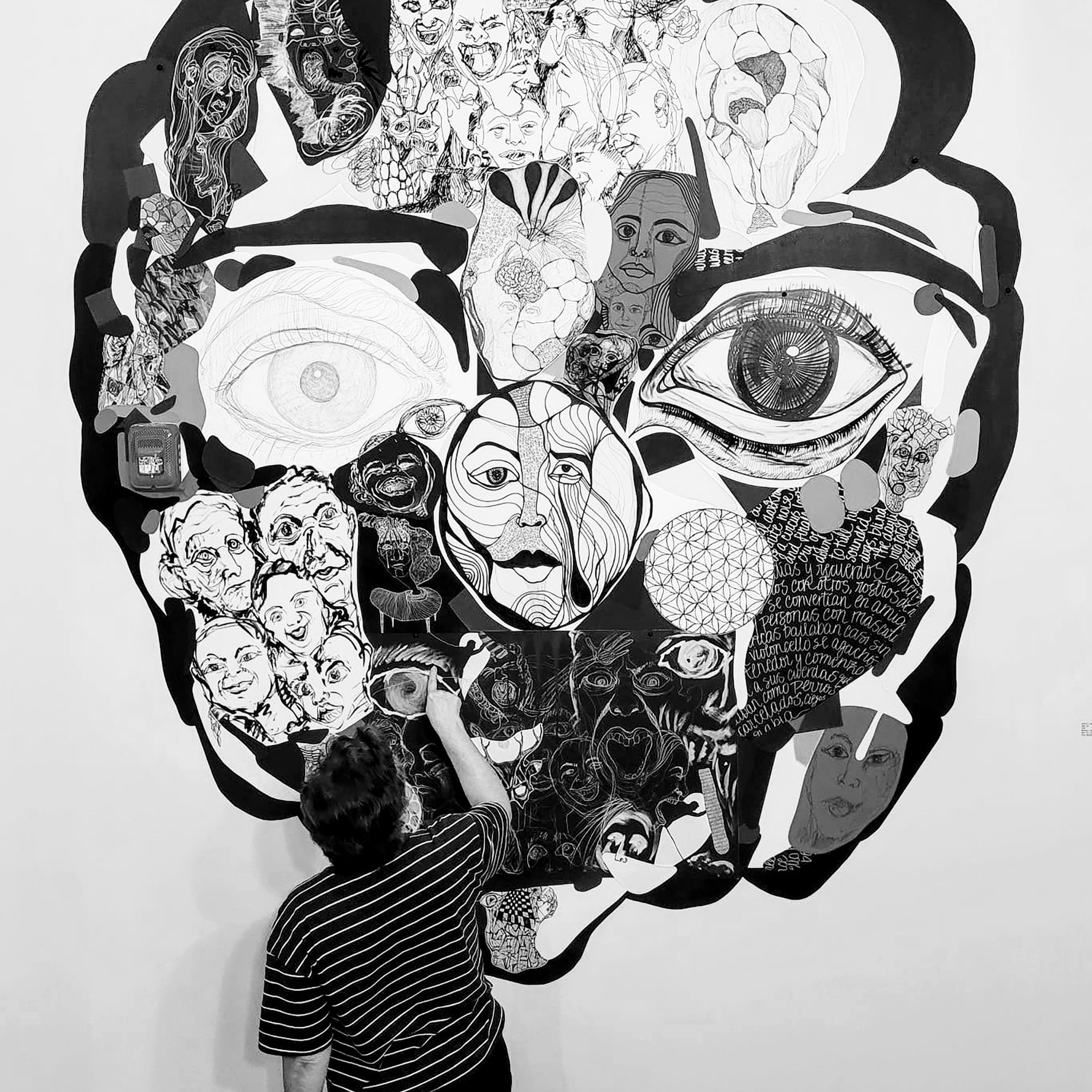 A black and white photo of Artist Gisela Romero as she draws on a mural of self portraits collaged into one large portrait.