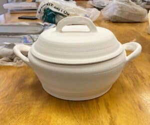 Unbaked white pot with lid and handles sitting on a yellow wooden table. 