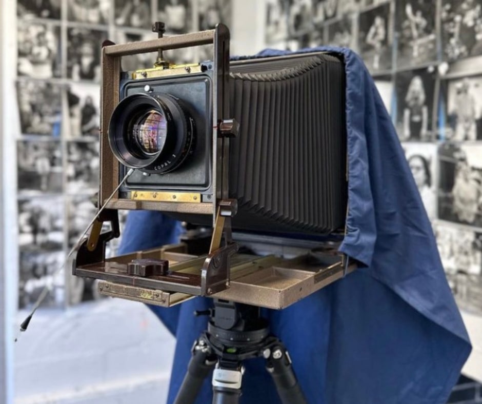 A vintage accordion style film camera in front of two walls filled with black and white portraits