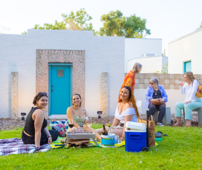 Three woman smiling at the camera while sitting on blankets having a picnics, 3 other people in the background having conversations.