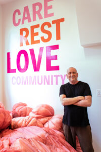 Chief Curator Dan L. Hess smiles at the camera while standing in front of wall text that says "Care, Rest, Love, and Community" by Jessica Caldas.
