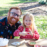 A father and his daughter smile happily at the cameras as they craft their nature pinch pot at one of our crafting tables in the main garden.