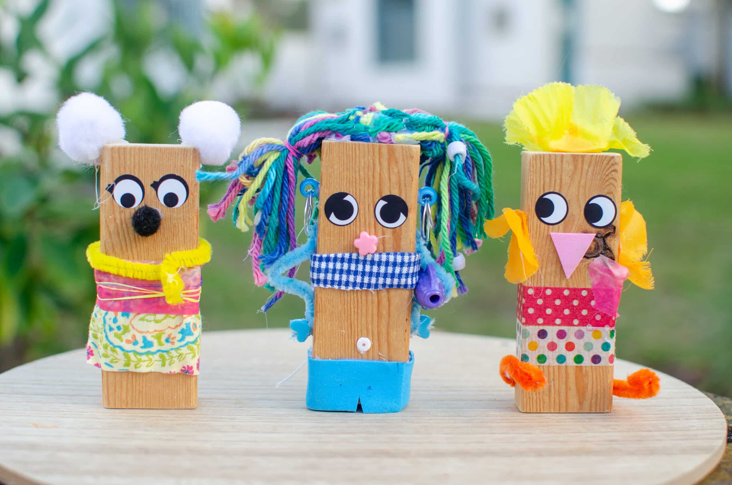 Group of three decorated wooden blocks with googly eyes, colorful pom poms, pipe cleaners, and ribbon.