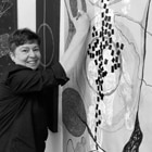 A Venezuelan woman with short, dark hair smiles while hold a drawing tool up against a giant composition.