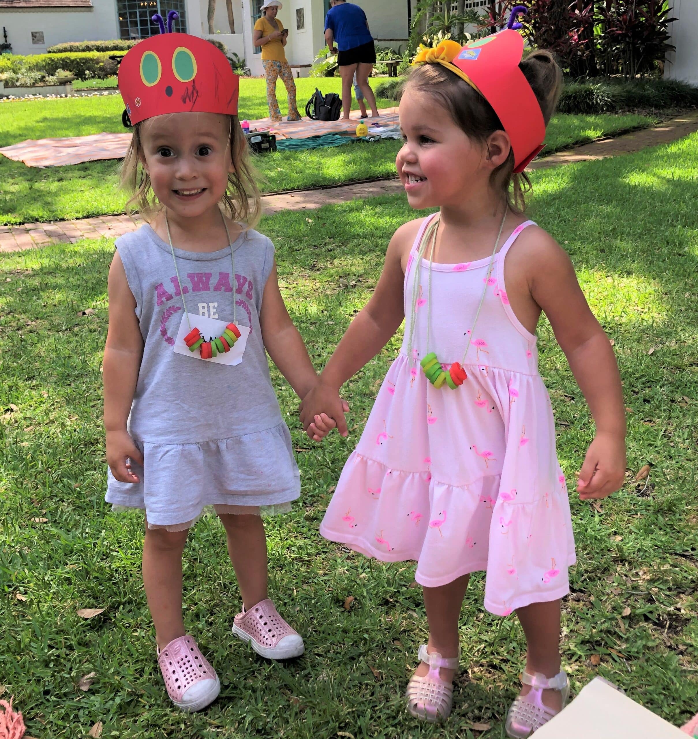 two young white toddlers wearing Hungry Caterpillar-themed craft crowns and necklaces hold hands and make excited faces in the A&H main garden