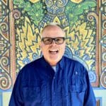 An older man with big black framed glasses, a bright blue shirt smiling in front of a mayan painting by Andre Smith.