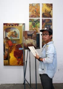 MJ, a Filipina with short black hair, tan skin, and dark eyes behind black rimmed glasses, stands with a sketchbook in front of an easel. She is wearing a ball cap and a long button down shirt spattered with paint. On the easel and the wall behind her hang her paintings, which are composed mostly of domestic or mundane scenes from daily life seen from a forced overhead perspective,