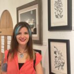 A woman with brown hair and blue streak framing her face smiling in front of a gallery wall.