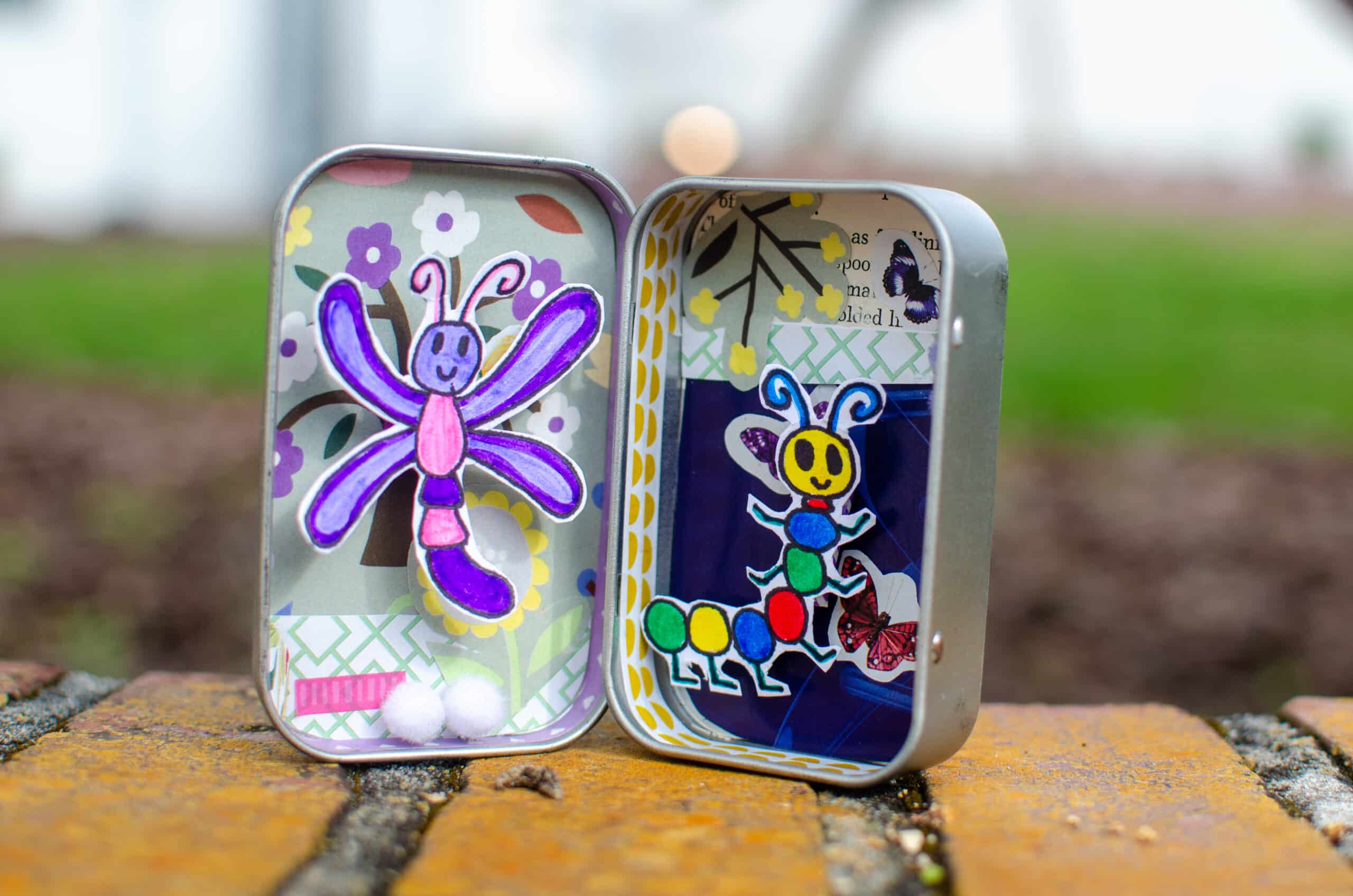 Tin box opened and decorated with paper, fabrics, and colorful drawings that represent bugs.
