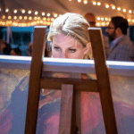 A blonde white lady with pretty blue eyes is looking at a canvas art piece that is leaning on a wooden easel in a lit-up outdoor event.