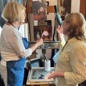 Patti and friend face away from the camera looking at a painting on an easel.