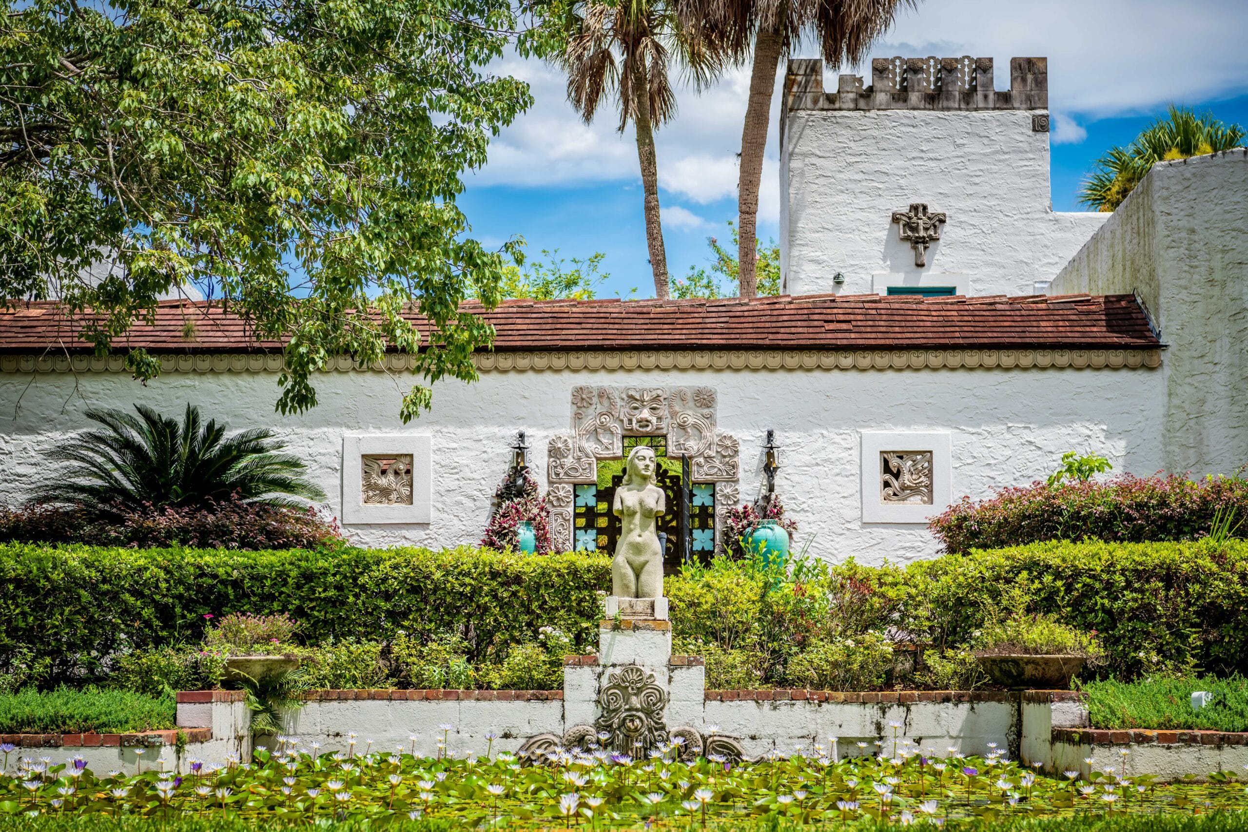 A pond full of blooming lilies is topped with a beautiful bust of Venus-style sculpture of a woman. She is framed by an intricately carved doorframe on a white stucco castle behind her. The castle is decorated with more carvings on its sides and atop its tall tower. Lush tropical plant life surrounds the scene.