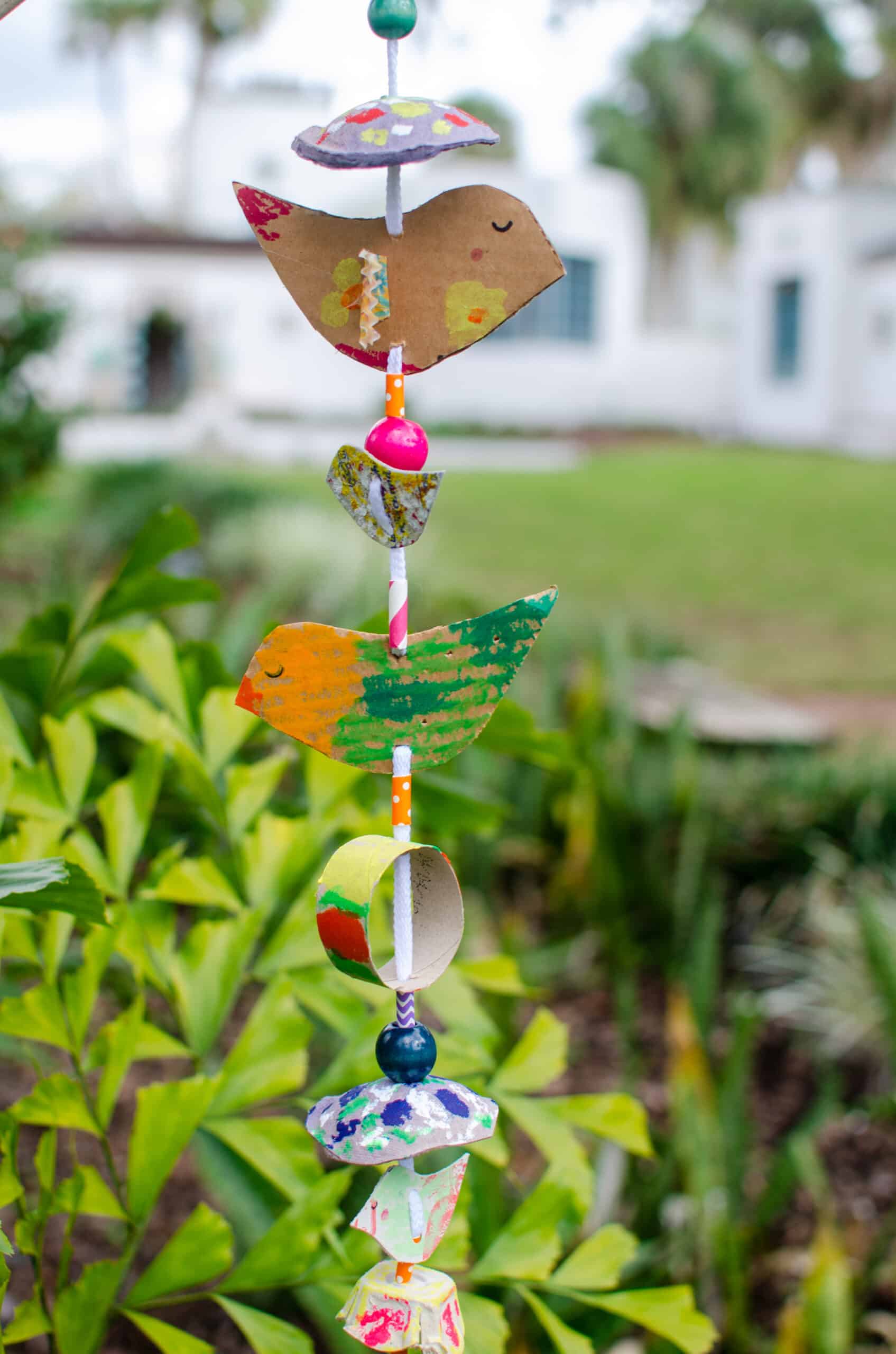 Colorful decoration that hangs vertically in front of vibrant bushes and leaves. Made of cardboard, beads, shoe laces, and paint. Decoration represents multiple cut-out cardboard birds, and other round organic shapes.