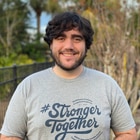 A young man smiles. He has short facial hair, and dark brown hair that goes to his ears. Trees and blue skies shimmer behind him.