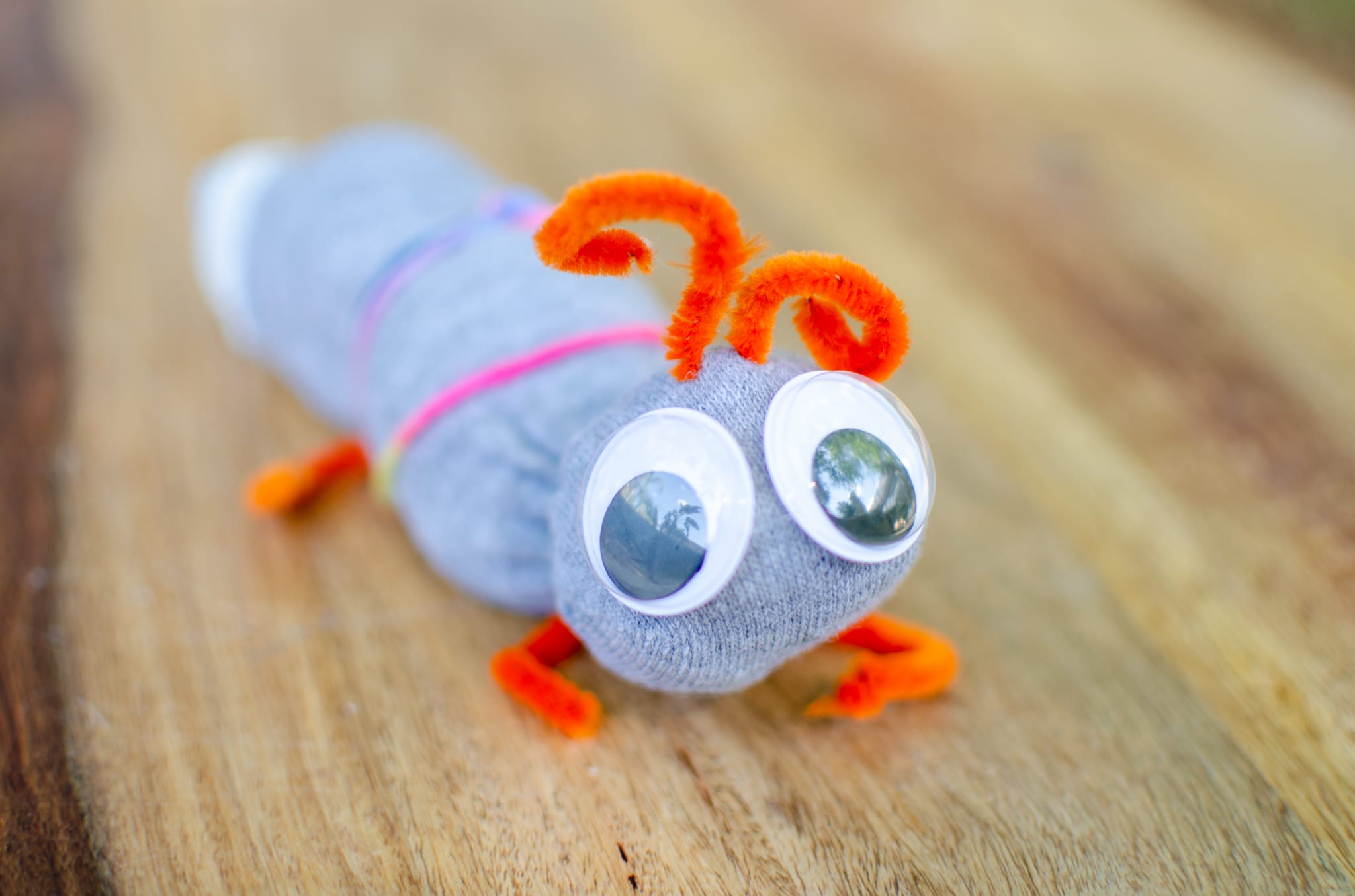 Image of a cute purple caterpillar that lays on a wooden table. Made of purple pom poms, orange pipe cleaners, and big googly eyes.