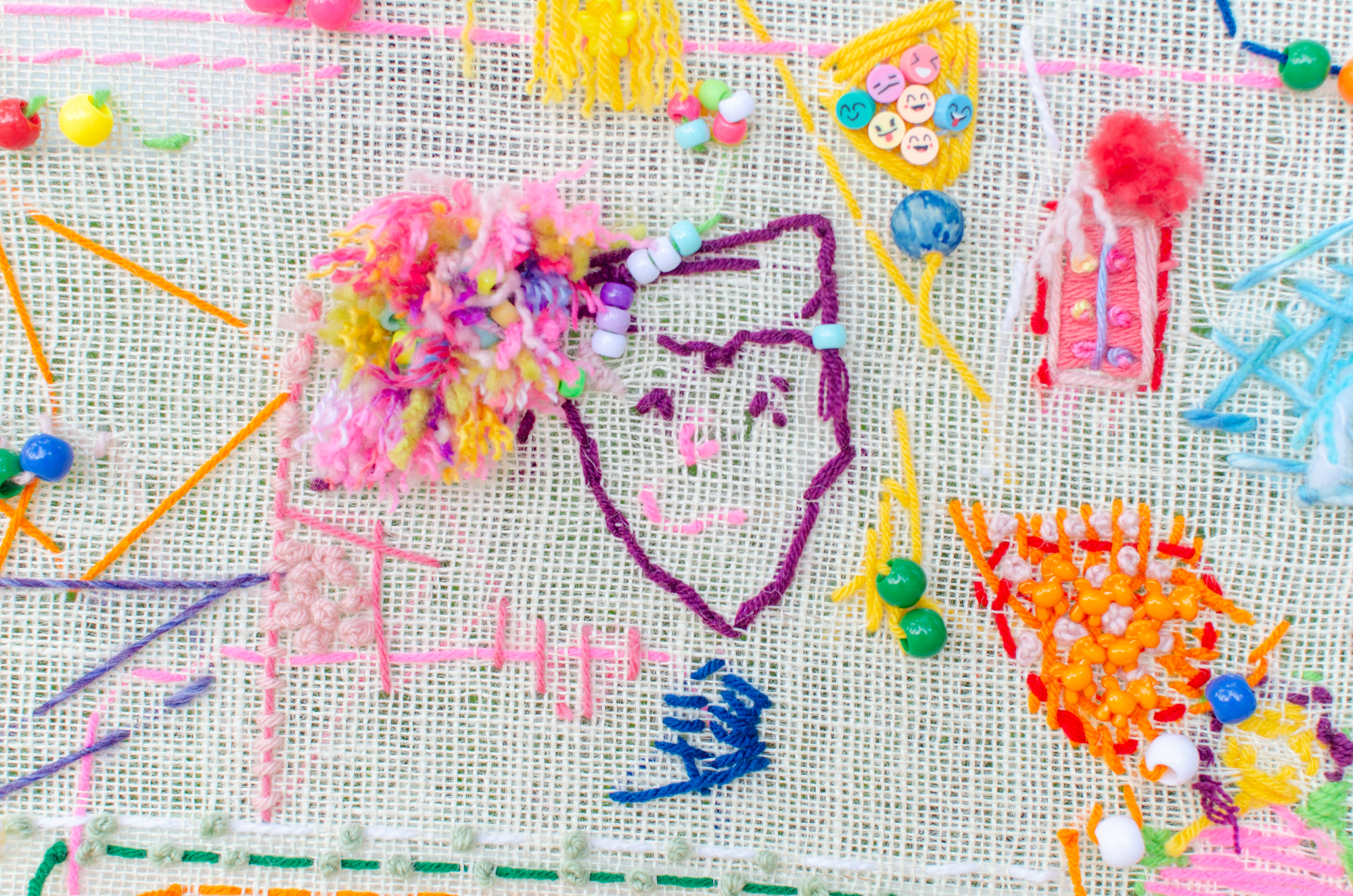 A close up image of a yarn tapestry with a smiling face in the center surrounded by other random objects in colorful yarn.