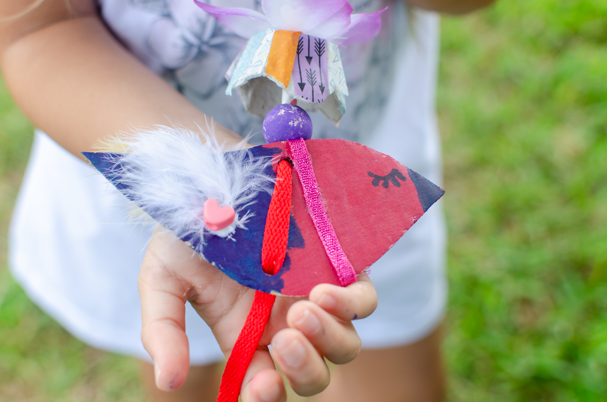 A young child holding their recycled bird sculpture made of cardboard, feathers, stickers, and a flower.