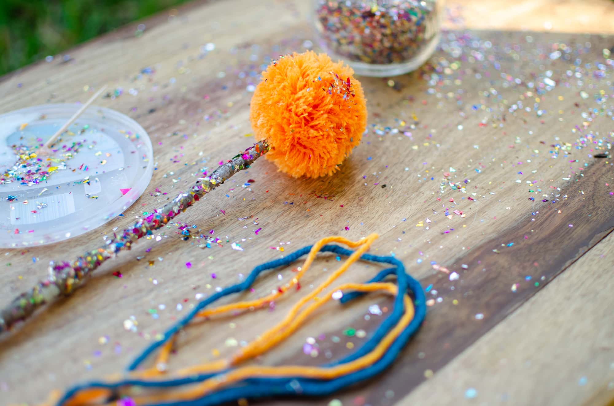 Colorful handmade stick with a bright orange pom pom on the top of a wooden table covered in colorful confetti.