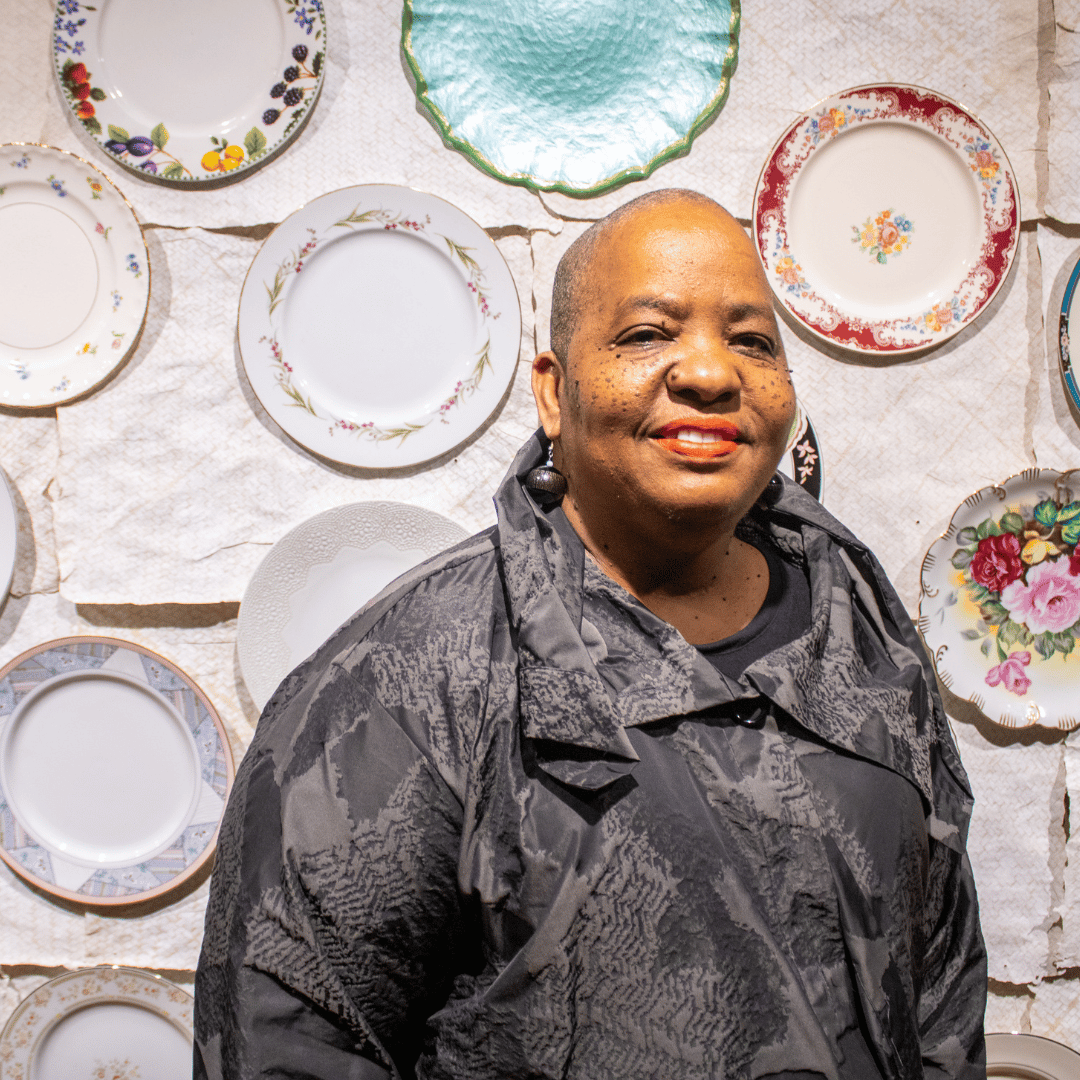 Kendra, a bald Black woman with freckles in a stylish black coat, stands smiling in front of her installation "Collecting Plates" at the Art & History Museums of Maitland's contemporary art gallery