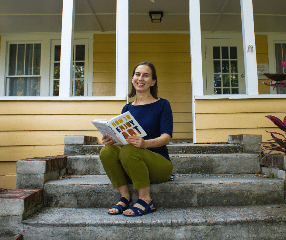 Jessi, a white woman with short brown hair, smiles with a book, seated on the steps of the charming yellow Cottage at Lake Lily