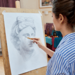 Woman in a striped shit using a pencil to draw a face on paper sitting on an easel.