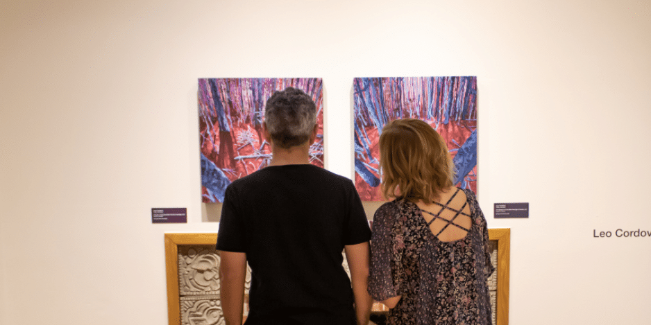 A couple standing in front of a fire place looking at two paintings that are inside the A&H Gallery.