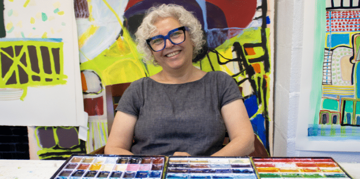 Carole, a white woman in her 60s with short curly white hair and large blue-framed glasses, sits smiling in a studio. In front of her are a colorful palette of paints. Behind her, one of her vibrant abstract works is visible.