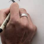 A close-up view of a light-skinned hand holding a drawing pen over a piece of paper. The hand has a silver ring on its middle finger.