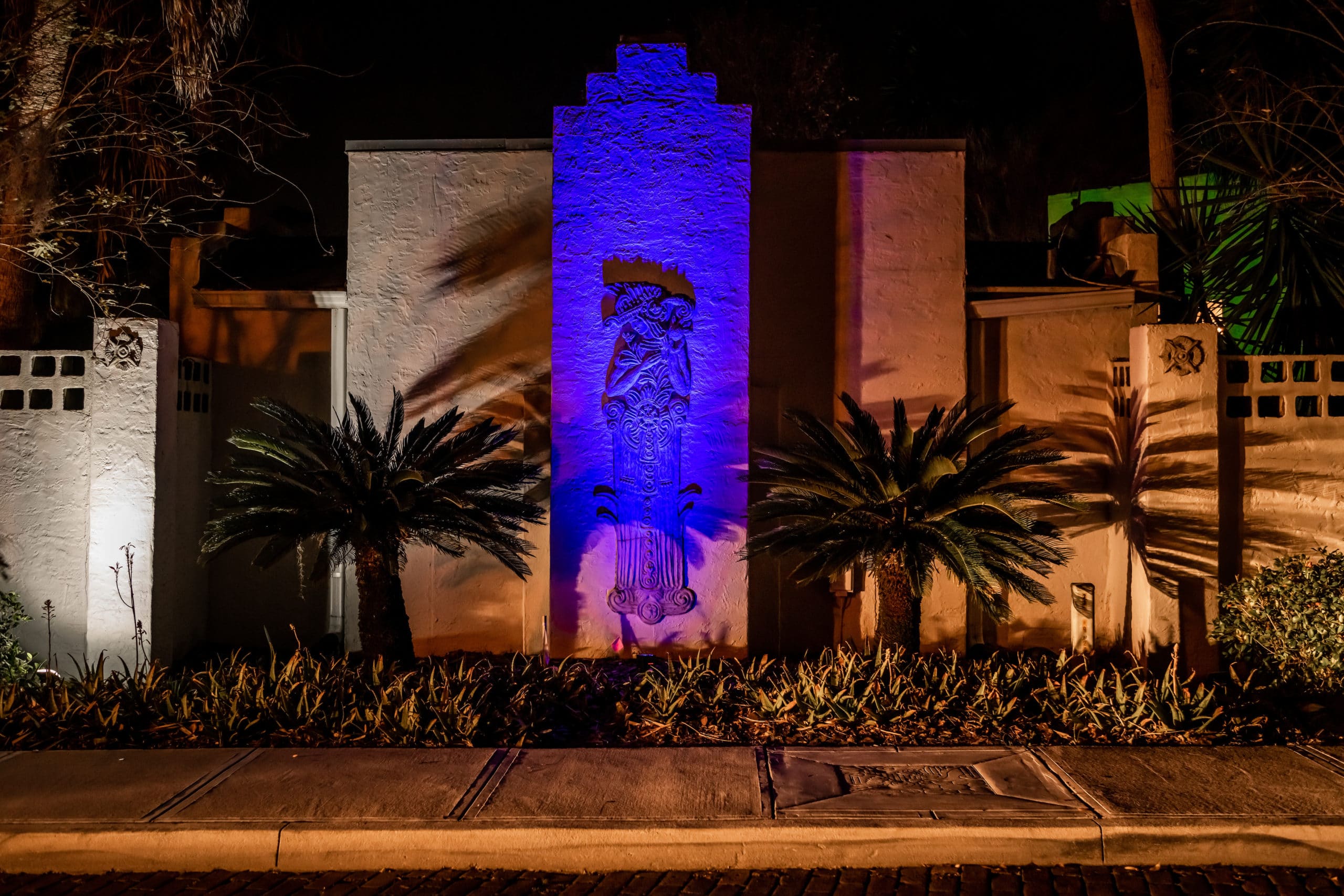 The outside of an A&H building with a large concrete relief sculpture of a person, imaginatively dressed in a complex costume reminiscent of old Mayan illustrations, dramatically and colorfully lit in the night.