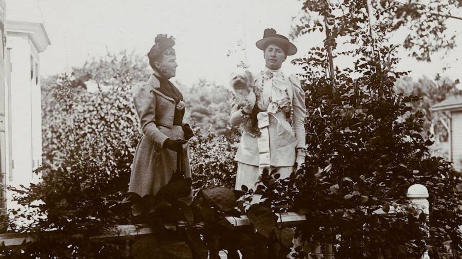 Vintage sepia photo of two women standing next to each other, outside and wearing Victorian clothing.